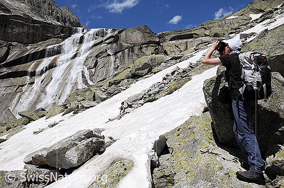Foto: Alpinistin vor Wasserfall. Das Wasser stürzt sich als breiter Wasserfall mit mehreren Armen über eine hohe Felswand. Im Vordergrund ist eine Geröllhalde mit Schneefeld zu sehen.