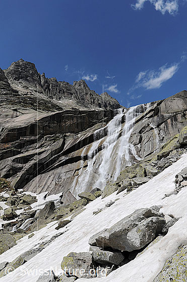 Foto: Gelmerhörner und hoher Wasserfall. Unterhalb der Wand liegt Geröll. Im Vordergrund ist ein Schneefeld zu sehen.