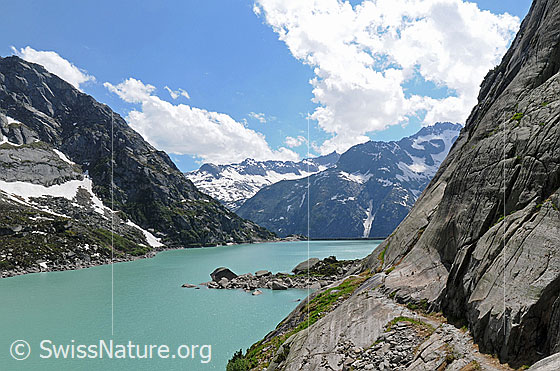 Foto: Bergweg am Gelmersee mit Blick über das türkisfarbene Wasser zur Staumauer. Über den Bergen (Juchlistock, Brünberg, Alplistock) steigen Quellwolken auf.
