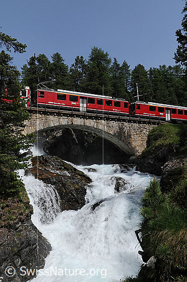 Foto: Eisenbahnbrücke über Bergbach mit tosendem Wasserfall. Über die Brücke fährt ein Personenzug der Rhätischen Bahn(RhB).