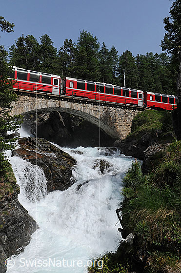 Foto: Rhätische Bahn (Unesco-Welterbe) auf Eisenbahnbrücke über Wasserfall im Engadin.
