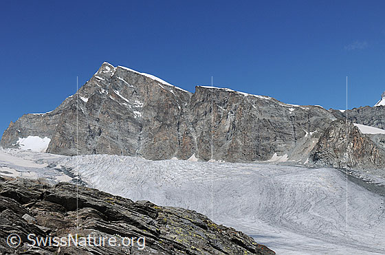 Foto: Allalinhorn und Allalingletscher. Die mächtigen Felswände erheben sich über dem Gletscher. Im Vordergrund ist eine Felsschichtung zu sehen.