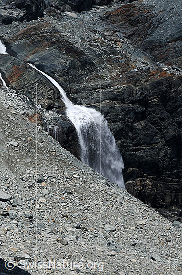 Foto: Wasserfall in karger Landschaft.
Im Fels eingefressener Bergbach und Wasserfall, welcher sich über eine Felsstufe stürzt. Die Umgebung ist karg und im Vordergrund ist ein Geröllfeld zu sehen.