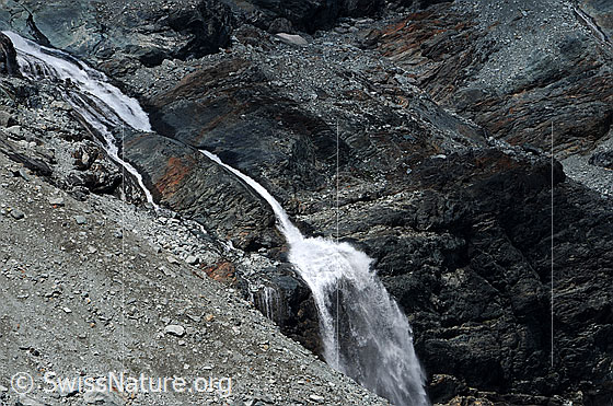 Foto: Geröll und Fels mit Wasserfall.
Im Fels eingefressener Bergbach und Wasserfall, welcher sich über eine Felsstufe stürzt. Die Umgebung ist karg und im Vordergrund ist ein Geröllfeld zu sehen.
