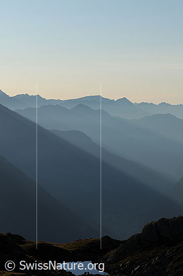 Foto: Morgenstimmung über der Leventina. Durch das schräg einfallende Morgenlicht sind die Bergketten, Berghänge und Gipfel (Vogelberg, Cima Rossa und Cima dei Cogn) als Silhouetten erkennbar. Im Vordergrund ein spiegelglatter Bergsee.