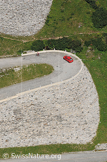 Foto: Haarnadelkurve des imposanten Bauwerks durch die Tremola. Ein Radfahrer und ein Auto befinden sich in der Kurve der Passstrasse.