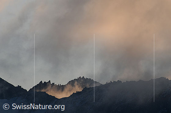 Foto: Morgenstimmung mit rötlichen Wolken und Streiflicht in Berglandschaft mit gezacktem Felsgrat.