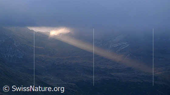 Foto: Ein Lichtstrahl bricht durch die Wolkendecke über dem Albrunpass und fällt auf die kalte, herbstliche Berglandschaft.