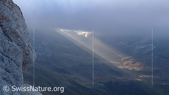 Foto: Morgenstimmung mit Lichtspot. Die Sonnenstrahlen brechen beim Albrunpass durch die Wolkendecke und beleuchten die urtümliche, herbstliche Berglandschaft mit Felsen im Vordergrund.
