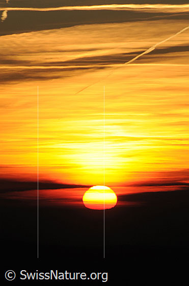 Foto: Sonnenuntergang. Die Sonne sinkt als helle Scheibe in eine Wolkenschicht. Der Himmel ist bewölkt und erscheint im Abendlicht in warmen Farbtönen zwischen rot und gelb.