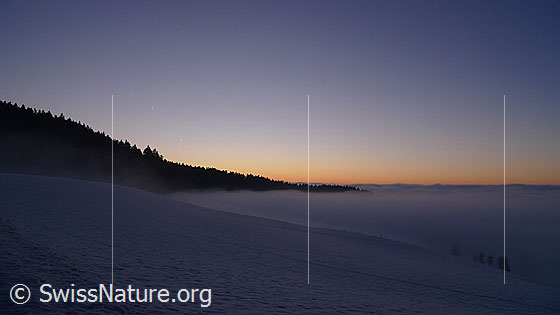 Foto: Rötlicher Abendhimmel am Horizont des umfassenden Nebelmeers. Das Nebelmeer grenzt an einen bewaldeten Hügelzug. Feine Nebelschleier ziehen sanft über die schneebedeckte Anhöhe an der Nebelgrenze.