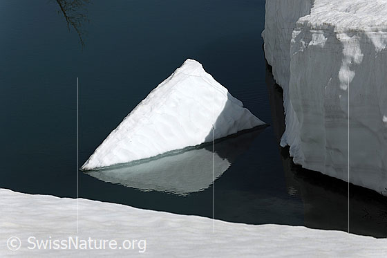 Foto: Eine leuchtend weisse Eisscholle mit der Form einer Pyramide treibt im Wasser und spiegelt sich darin. Das Ufer des Wasserlaufs wird durch eine kompakte Schneedecke begrenzt.