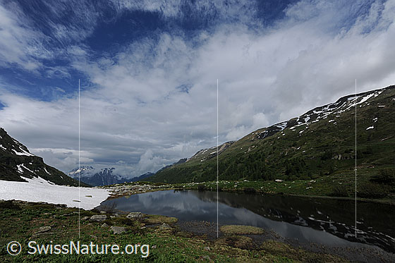 Foto: Aufreissende Wolkendecke über dem Halsesee. Im ruhigen Bergsee spiegelt sich ein Bergrücken und am Ufer ist ein Schneefeld zu sehen.