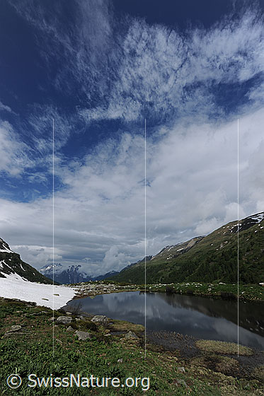 Foto: Aufreissende Wolkendecke über dem Halsesee. Im ruhigen Bergsee spiegelt sich ein Bergrücken und am Ufer ist ein Schneefeld zu sehen.