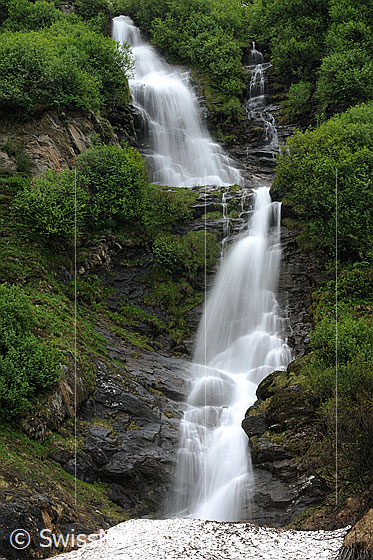 Foto: Wasserfall als Langzeitaufnahme. Das Wasser fliesst über gestufte Felsen, welche mit Sträuchern in hellem Grün bewachsen sind.