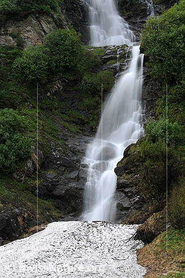 Foto: Mit Sträuchern bewachsener Berghang mit mehreren Felsstufen, über welche sich ein Wasserfall ergiesst (Langzeitaufnahme). Im Vordergrund ist ein Schneefeld zu sehen.