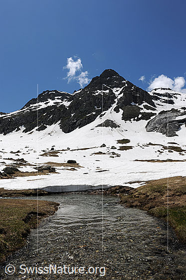 Foto: Berge und Wasserlauf. Klares Wasser eines ruhigen Bergbachs fliesst über die Hochebene, welche teilweise noch schneebedeckt ist. Über den Berggipfeln im Hintergrund steigen erste Quellwolken auf.