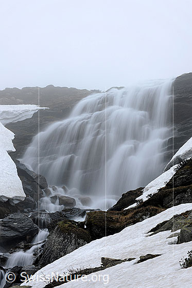 Foto: Fliessendes Wasser. In feinen Strahlen fliesst das Wasser eines Wasserfalls über einen leicht gestuften Felsriegel. Im Bachbett liegen Felsblöcke und in der Umgebung sind noch Schneefelder zu sehen.
