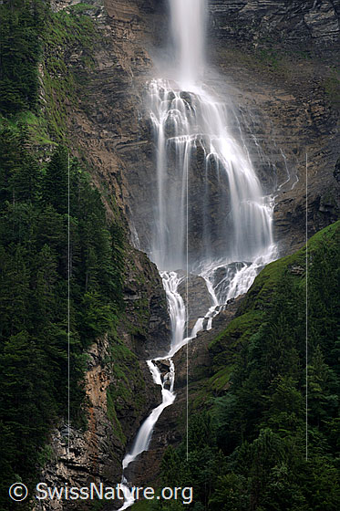 Foto: Ein Wasserfall trifft auf einen Felsvorsprung, ergiesst sich darüber und fliesst als Bergbach durch bewaldetes Gebiet weiter talwärts.