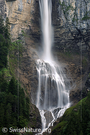 Foto: Ein Wasserfall trifft auf einen Felsvorsprung in einer Felswand, ergiesst sich darüber und fliesst als Bergbach durch bewaldetes Gebiet weiter talwärts.