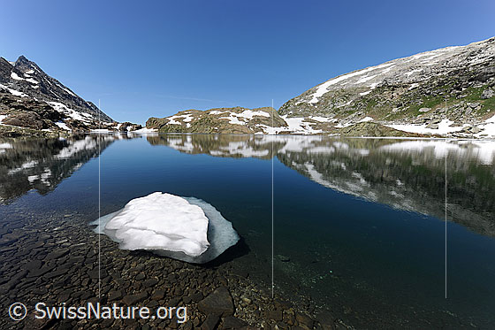 Foto: Berglandschaft mit Schneeresten und Spiegelung im Geisspfadsee. Am Ufer des Bergsees sind eine kleine Eisscholle und flache Steine zu sehen.