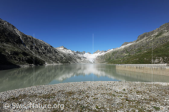 Foto: Oberaarsee und Teile eines Speicherkraftwerks. Im Stausee spiegeln sich Staumauer und Berglandschaft mit Oberaargletscher und Oberaarhorn.