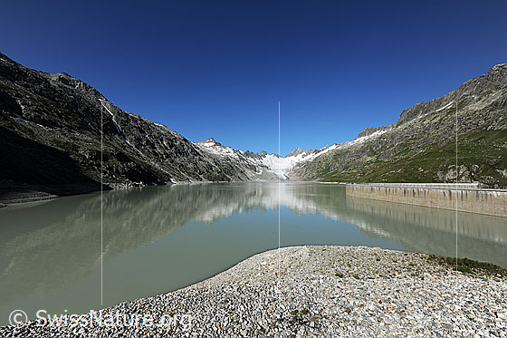 Foto: Teile eines Speicherpumpwerks am Oberaarsee. Staumauer und Berglandschaft spiegeln sich in der Wasserfläche des Stausees.