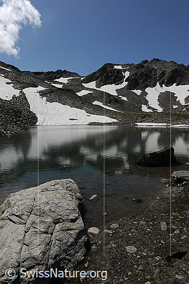 Foto: Spiegelung im Schwärziseeli (Mittlerer Bergsee). Am Ufer befinden sich Felsblöcke und in der Berglandschaft im Hintergrund liegen noch Schneereste.