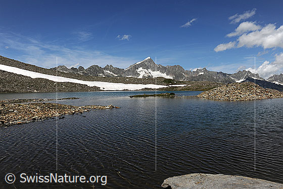 Foto: Schwärziseeli (Oberster Bergsee) mit Wellen und Kiesbänken. Ein Schneefeld reicht ans Ufer. Hintergrund: Bergkette mit Galenstock und Quellwolken.