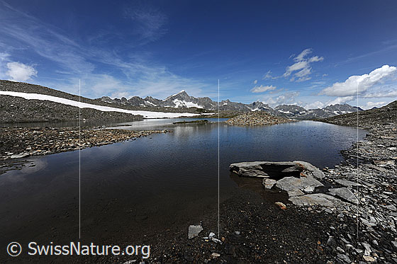 Foto: Seelandschaft Schwärziseeli (Oberster Bergsee) mit Felsplatte am Ufer. Die Wasserfläche wird durch Kiesbänke unterbrochen. Über der Bergkette mit Galenstock sind Schleierwolken und Quellwolken zu sehen.