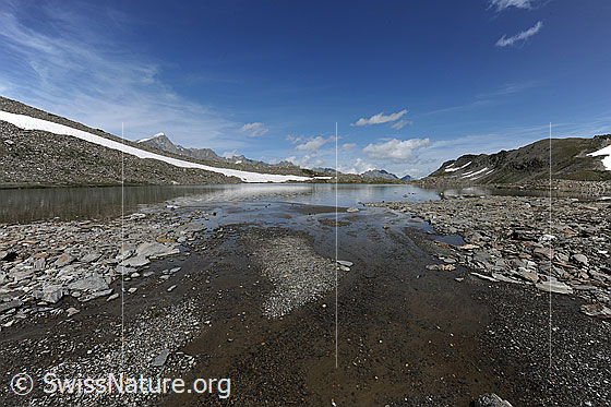 Foto: Schwemmlandschaft und Schwärziseeli (Oberster Bergsee). Das flache Ufer besteht aus kleinen Inseln aus Kies und Steinplatten. Über der Bergkette sind Schleierwolken und kleine Quellwolken zu sehen.