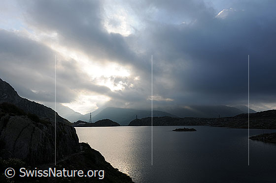 Foto: Stimmung über dem Totesee, Grimselpass. Wolkenstimmung der ausgedehnten Restbewölkung über der Seelandschaft. In den Felsen dem Bergsee entlang verläuft ein Bergweg.