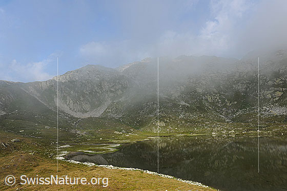Foto: Jostsee mit ruhiger Wasserfläche in welcher sich ein Berghang spiegelt. Der Bergsee ist von Alpweiden umgeben und die Restbewölkung löst sich langsam auf.