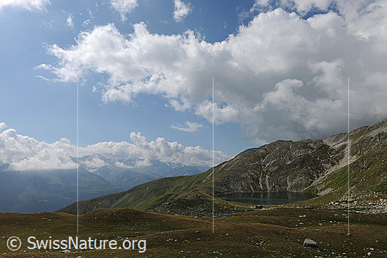 Foto: Rundsee in lieblicher Berglandschaft mit Alpweiden. Über den Bergen liegt ausgedehnte Quellbewölkung.