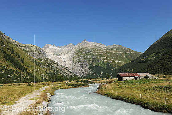 Foto: Gletschbode und Rotte (Fluss Rhone) mit Wanderweg dem Fluss entlang, Alpweiden und einem Stall. Im Hintergrund sind das Klein Furkahorn und die Passstrasse zum Furkapass zu sehen.