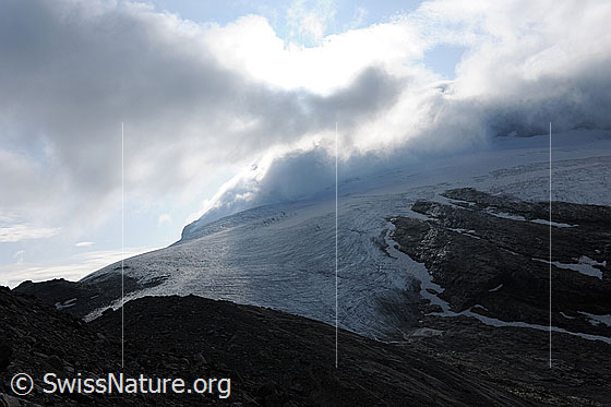 Foto: Wolkenstimmung über dem Chaltwassergletscher. Die Wolken hängen tief über dem Gletscher. Am Gletscherrand wurden durch das Abschmelzen Felsbänder freigelegt.