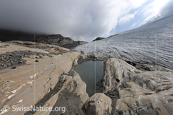 Foto: Dramatische Wolkenstimmung über Gletscherlandschaft. Vom Gletscher abgeschliffene Felsen mit feiner Zeichnung, Mustern und interessanten Verformungen, in welchen sich Wasser angesammelt hat. Auf dem Gletscherschliff wurden Steine und Sand abgelagert. Der Chaltwassergletscher hat an seinem Rand nur noch eine dünne Eisschicht.