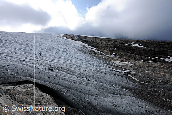 Foto: Chaltwassergletscher, Simplon. Flach in Fels und Geröll auslaufender Gletscher mit hellen und dunklen Wolken über der Gratkante. Am Gletscherrand im Vordergrund ist ein Hohlraum (Randkluft) zwischen Gletschereis und Fels zu sehen.