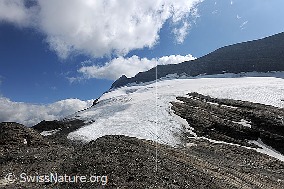 Foto: Chaltwassergletscher und Felsgrat am Monte Leone. Über der Berglandschaft mit Gletscher zieren Quellwolken den blauen Himmel. Am Gletscherrand sind kompakte Felsen und Geröllfelder zu sehen.