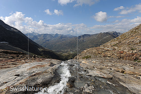 Foto: Simplongebiet. Bergbach in felsiger Landschaft mit Ausblick auf den Simplonpass und die Berglandschaft mit Straffelgrat und Tochuhorn. Darüber ist Quellbewölkung zu sehen. Die Felsen im Vordergrund sind vom Wasser abgeschliffen.