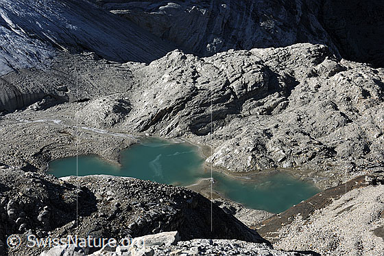 Foto: See im Vorfeld des Brunegggletschers. Im Gletschersee sind Eisreste zu sehen. Die Umgebung mit Felsen und Gletscherzunge ist mit feinem Geröll bedeckt. Licht und Schatten liegen auf der Gletscherlandschaft.