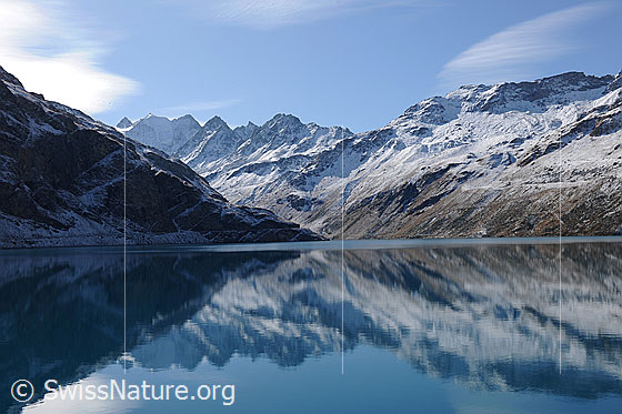 Foto: Spiegelung im Lac de Moiry. Das hellblaue Wasser des Stausees liegt ruhig in der herbstlichen Landschaft mit verschneiter Bergkette.