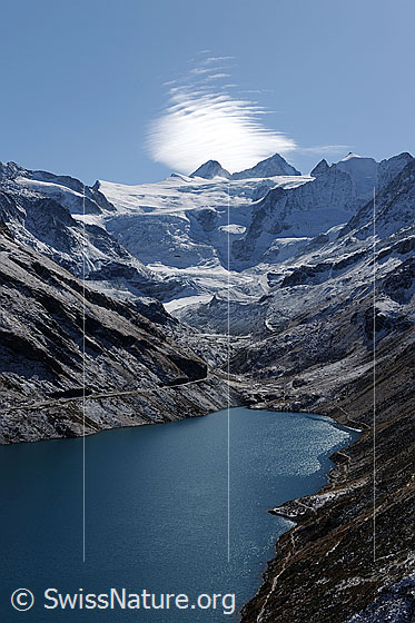 Foto: Lac de Moiry und Glacier de Moiry. Über den Gipfeln von Grand Cornier und Dent Blanche ist eine Föhnwolke zu sehen. Die Berglandschaft um den Stausee mit blau schimmerndem Wasser ist leicht verschneit.
