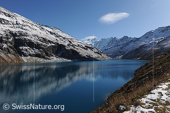 Foto: Lac de Moiry mit schwacher Spiegelung der Berge im blauen Wasser. Die Alpweiden sind herbstlich gefärbt und es liegt erster Schnee.
