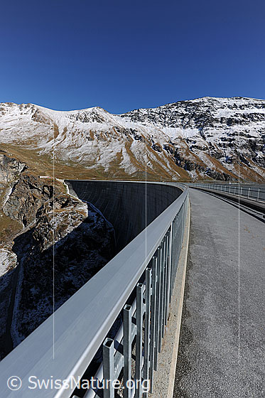 Foto: Am Geländer der Bogenstaumauer des Lac de Moiry. Blick über die Krone der Staumauer auf die herbstliche, leicht verschneite Berglandschaft.