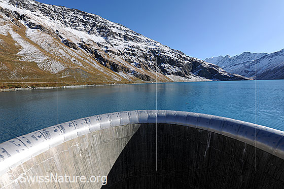 Foto: Überlauf an der Staumauer des Lac de Moiry. Blick über das blaue Wasser in die leicht verschneite Berglandschaft.