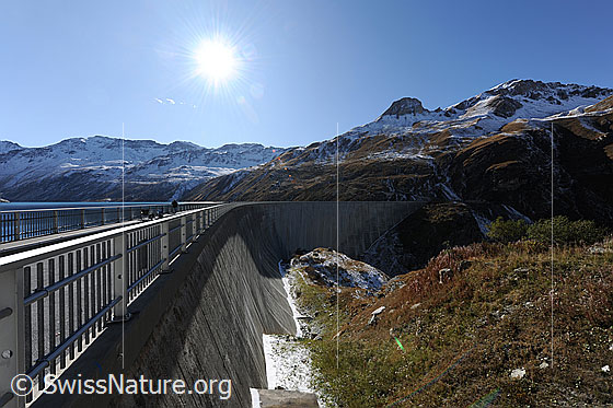 Foto: Staumauer des Lac de Moiry mit massivem Geländer. Erster Schnee liegt in der herbstlich gefärbten Berglandschaft. Darüber ist die Sonne am blauen Himmel zu sehen.