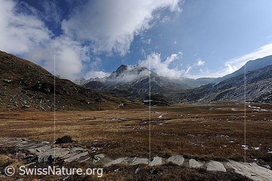 Foto: Die Weite eines herbstlichen Hochtals. Im Vordergrund verläuft ein Römerweg (Wanderweg) über die Ebene. Über dem Ofenhorn reissen die Quellwolken auf.