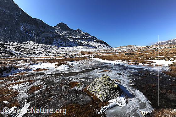 Foto: Gefrorener Wasserlauf in herbstlicher Hochebene. Teile des flachen Gewässers sind mit einer Eisschicht bedeckt. Die Berglandschaft ist leicht mit erstem Schnee überzuckert.