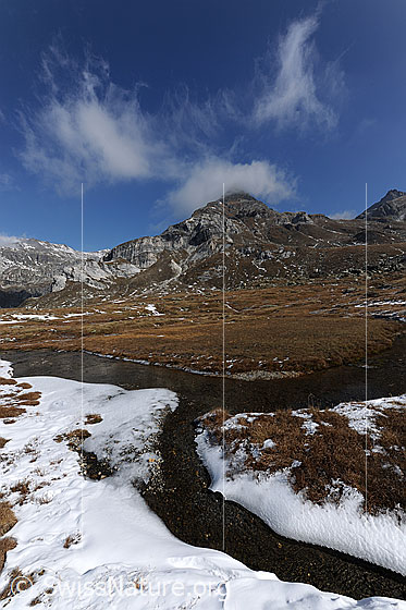 Foto: Wasserläufe in herbstlicher Hochebene. Im Vordergrund liegen noch Reste des ersten Schnees. Über dem Berggipfel im Hintergrund sind Wolkenfetzen zu sehen.